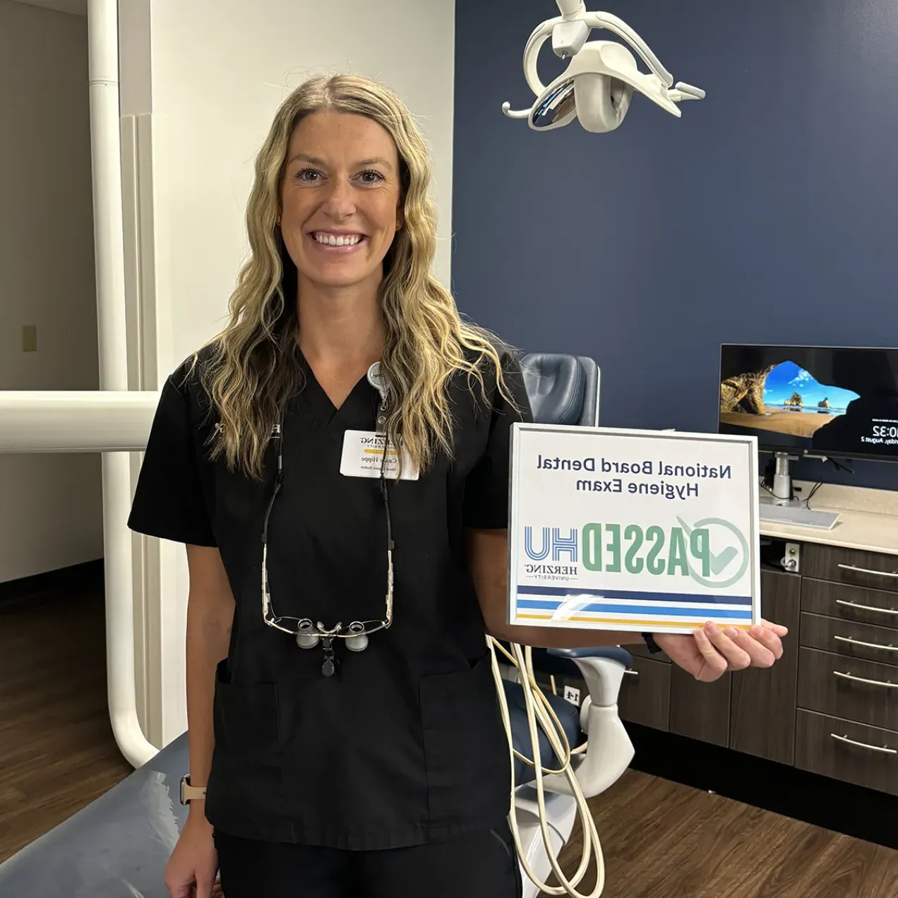 A female dental hygiene student in black scrubs smiles while holding a ‘Passed National Board Dental Hygiene Exam’ certificate in a dental clinic setting, with a Herzing University ID badge visible.